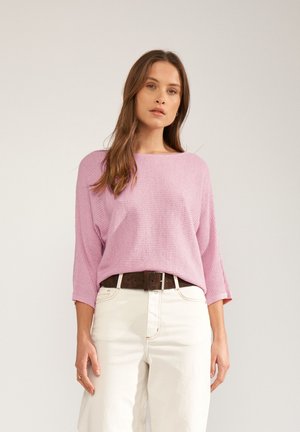 Nő hosszú barna hajjal, laza rózsaszín kötött pulóvert, fehér magasderekú nadrágot és sötétbarna övet visel, egyszerű háttér előtt áll.