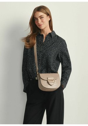 Kobieta o długich brązowych włosach, ubrana w czarną koszulę w groszki, czarne spodnie oraz beżową torebkę na pasku z złotą klamrą.