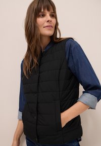 Gilet imbottito nero con colletto alto, dotato di chiusure a bottoni e tasche laterali, indossato sopra una camicia di denim. Texture liscia, design leggero.