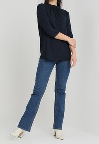 Blusa azul marino con escote fruncido, mangas de tres cuartos, combinada con jeans de mezclilla azul de campana y botas de tacón blanco.