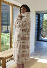 Płaszcz polarowy z dużym kołnierzem, w kremowym kolorze z beżowym wzorem w kratę, z bocznymi kieszeniami i luźnym krojem, umieszczony w jasnej przestrzeni wewnętrznej.