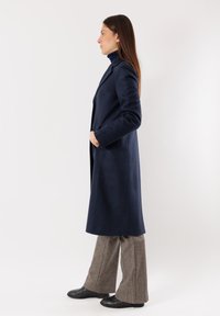Piumino lungo in misto lana blu navy con taglio dritto, rever a punta, tasche laterali e tessuto testurizzato, indossato sopra pantaloni larghi a fantasia.