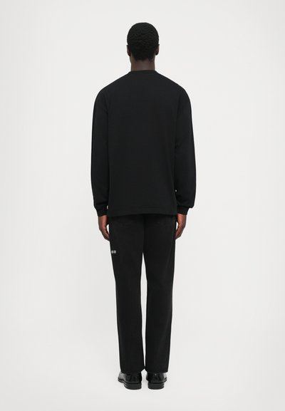 Mann steht mit dem Rücken zur Kamera und trägt ein schwarzes Langarmshirt, schwarze Hosen mit einem kleinen weißen Patch am rechten Bein und schwarze Schuhe.