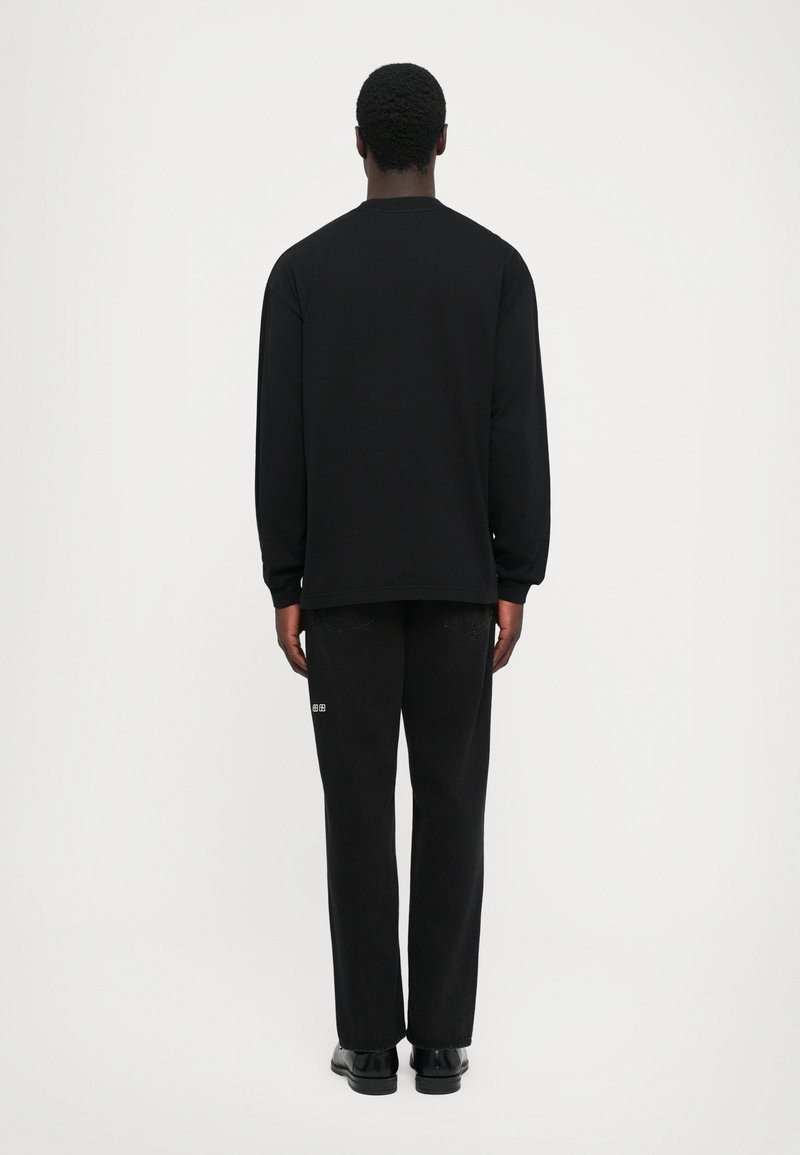 Homme de dos portant une chemise à manches longues noire, un pantalon noir avec une petite pièce blanche sur la jambe droite, et des chaussures noires.