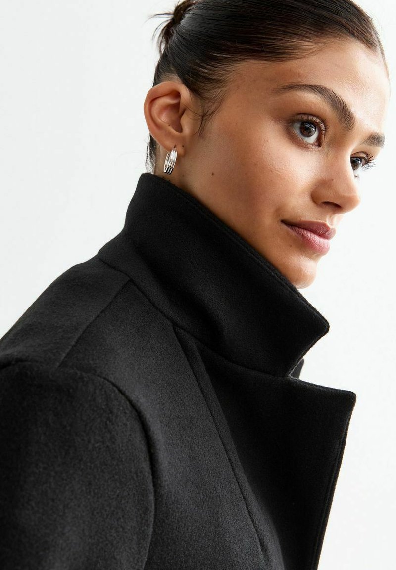 Manteau en laine noir avec un col montant, un tissu texturé et des revers structurés. Le mannequin porte des boucles d'oreilles en argent et a une coiffure soignée.