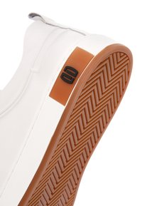 Biała wsuwana but z brązową gumową podeszwą z zygzakowatym bieżnikiem oraz małym czarnym logo na boku w pobliżu pięty.