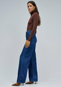 Camiseta marrón de manga larga y cuello alto combinada con un jeans azul de tiro alto y pierna ancha. Acentuada con detalles de botones en los puños.