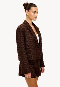 Blazer texturé marron avec des motifs en vagues, shorts assortis et col décontracté. Le matériau semble doux et moelleux avec une finition légèrement brillante.