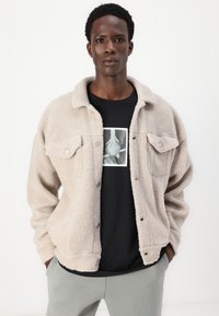 Beige fleece jack met drukknoopjes, twee borstzakken, gedragen over een zwart t-shirt met een monochroom ontwerp. Grijze broek is ook zichtbaar.