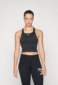 Svart sports crop-top laget av elastisk materiale med brede stropper og rund halslinning. Kombinert med svarte leggings med en hvit logo.