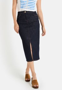 Falda lápiz de mezclilla oscura con una abertura frontal, terminada con costuras en contraste. Combinada con una blusa blanca y zapatos de slip on marrones.