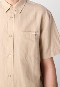 Beiges kurzärmliges Hemd mit Knopfleiste und einer Brusttasche vorne, die ein Calvin Klein-Label auf dem oberen Oberkörper einer Person zeigt.