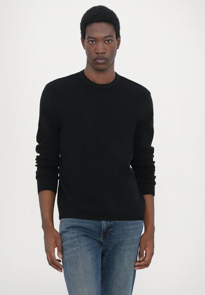 Pull à côtes noir avec un col rond et des manches longues, associé à un jean bleu. Le pull présente un tissu texturé avec une coupe ajustée.