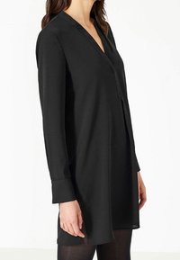 Czarna tunika z długim rękawem i dekoltem w serek, o prostym kroju i gładkiej teksturze, sięgająca do połowy uda, z zapięciem na guziki przy mankietach.