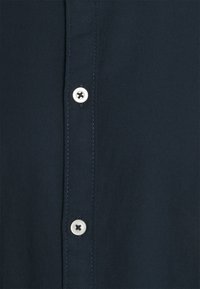 Marineblaues Hemd mit Knopfleiste und glatter Textur, das einen weißen Knopf mit einem Kreuzdesign und kontrastierenden Nähten entlang der Knopfleiste aufweist.