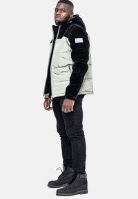 Vadderad jacka med en ljusgrå framdel och svarta plyschärmar, med dragkedjestängning, huva och logopatch på ärmen. Svarta byxor och stövlar.