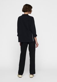 Zwarte langemouwen top met opgerolde manchet en zijnaad, gecombineerd met losse zwarte broek. Kenmerkt zich door een minimalistisch, ontspannen ontwerp.
