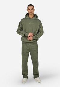 Sudadera verde oliva con puños y cinturilla acanalados, que presenta el logo "PEGADOR". Combinada con pantalones acanalados a juego y zapatillas blancas.