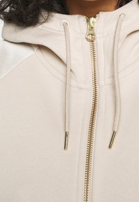 Beige zip-up hoodie med en gyllene dragkedja, snoddar och texturerade axeldetaljer. Mjuk tyg med en avslappnad passform.