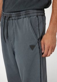 Pilkos joggingo kelnės, pagamintos iš lengvos medžiagos, su elastinga juosta, užtrauktuku, šoninėmis kišenėmis ir trikampiu logotipo aplikacija.