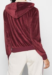 Sudadera de terciopelo burdeos con una textura suave, que cuenta con capucha, mangas largas y un dobladillo acanalado, combinada con pantalones de color claro.