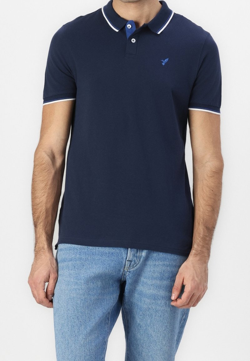 Homme portant un polo bleu marine à manches courtes avec des bordures blanches et un jean bleu clair, debout devant un fond uni.