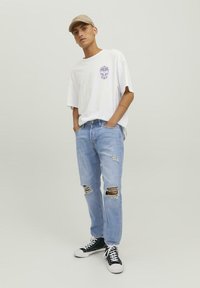 Camiseta blanca de gran tamaño con gráfico morado, jeans desgastados de corte claro con rotos en las rodillas, y zapatillas negras de caña alta. Lleva una gorra beige.