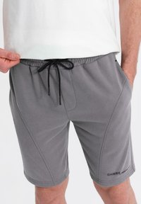 Shorts de sport gris en tissu doux, dotés d'une taille élastique avec cordon de serrage, de poches latérales et d'un logo noir sur la jambe gauche.
