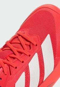 Červená sportovní bota s texturovaným síťovaným svrškem, plochými tkaničkami a stříbrným designem s třemi pruhy. Kulatá špička a polstrovaná podrážka.