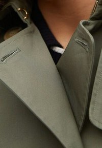 Zielony płaszcz o teksturowanej tkaninie, z zaostrzonymi klapami, widocznymi szwami oraz subtelnymi akcentami metalowymi w okolicy kołnierza.