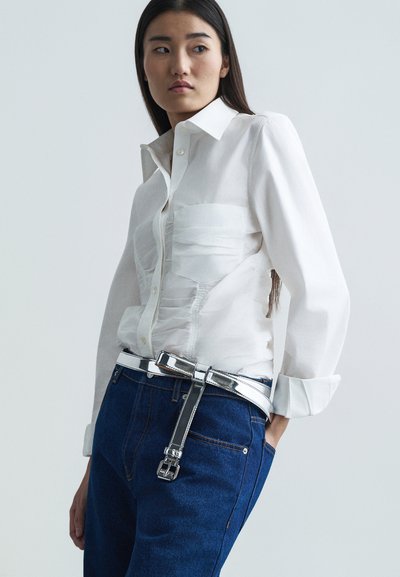 Λευκό πουκάμισο με υφή στο μπροστινό μέρος, σε συνδυασμό με σκούρο μπλε τζιν και μια γυαλιστερή ασημένια ζώνη με μεταλλική αγκράφα.