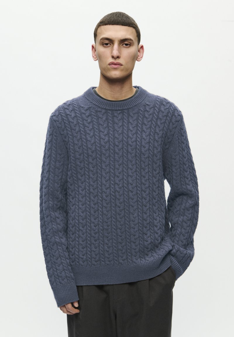 Modrý pletený svetr s texturovaným kabelovým vzorem, kulatým výstřihem a dlouhými rukávy. Ribované lemy a manžety. Nošený přes tmavé kalhoty.