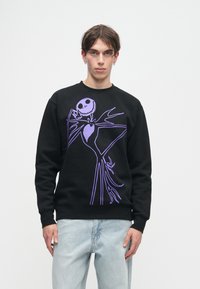 Jovem vestido com um sweatshirt preto com uma grande ilustração de um esqueleto roxo e umas calças de ganga azul claro, posando contra um fundo branco simples.