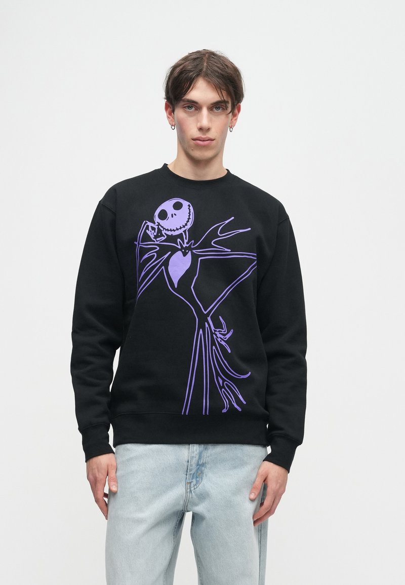 Jovem vestido com um sweatshirt preto com uma grande ilustração de um esqueleto roxo e umas calças de ganga azul claro, posando contra um fundo branco simples.