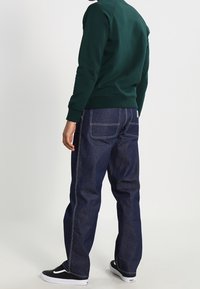 Mörkblå denimjeans med avslappnad passform, synliga sömmar och en klassisk femficksdesign. Svarta sneakers med en vit swoosh.