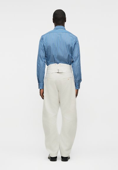 Άνδρας που φαίνεται από πίσω, φοράει μπλε ριγέ μακρυμάνικο πουκάμισο, φορεμένο μέσα σε ψηλόμεσα λευκά παντελόνια με αγκράφα στην πλάτη.