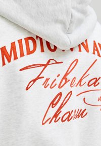 Gaišpelēka kapuce ar spilgtu oranžu izšūtu tekstu, kas apvieno dažādus drukas stilus uz muguras, mīksta tekstūra un kangoru kabata.