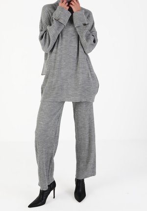 Femme portant un pull en tricot côtelé gris et un pantalon avec des bottes noires à talons hauts et bout pointu, debout sur fond blanc.