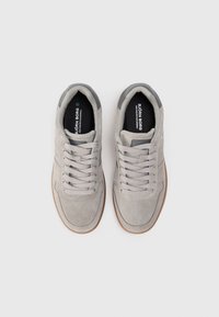 Zapatillas de ante gris claro con una textura suave, forma redondeada, acentos grises y una suela de goma marrón. Presenta seis ojetes y un logo en la lengüeta.