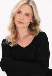 Pulover negru cu decolteu în V și mâneci lungi. Modelul are părul ondulat, blond și poartă cercei mici din argint, pe un fundal neutru.