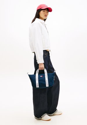 Námořnická látková taška s bílými popruhy a logem, držená osobou v bílé bundě a tmavých džínách, na jednobarevném pozadí.