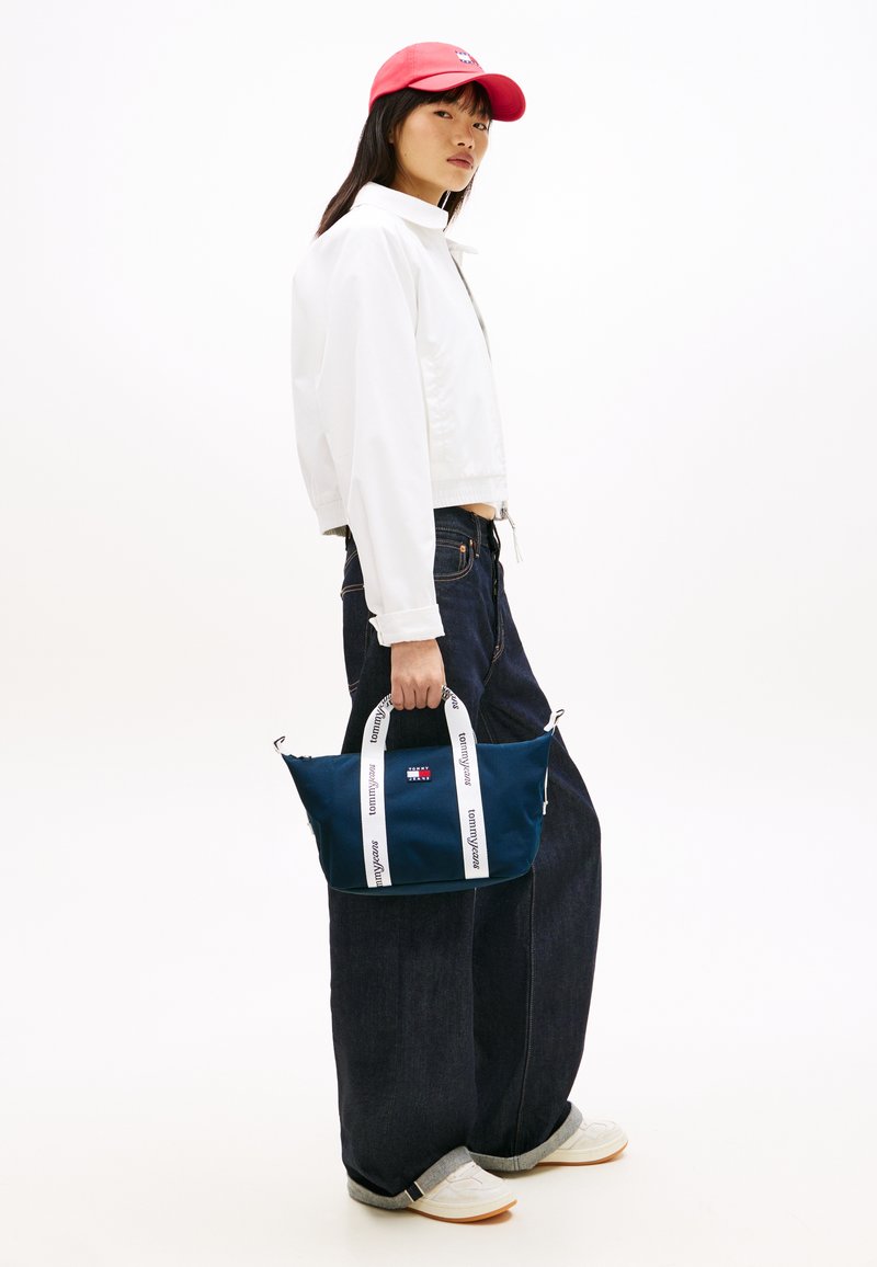 Τσάντα από ναυτικό ύφασμα με λευ κές τιράντες που φέρουν λογότυπο, κρατιέται από ένα άτομο με λευκό μπουφάν και σκούρο τζιν, σε απλό φόντο.