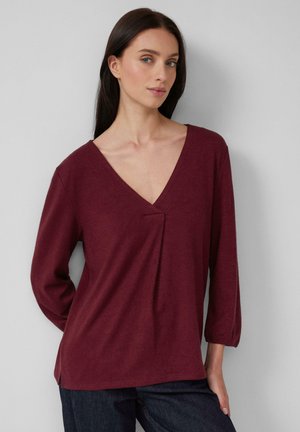 Donna con lunghi capelli scuri che indossa una maglia bordeaux a V con dettagli plissettati e jeans scuri, in piedi contro un muro grigio chiaro.