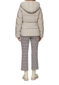 Dikke beige jas met een capuchon, voorzien van gestikte secties. Gecombineerd met gemusterde, cropped broeken in wit, rood en zwart.