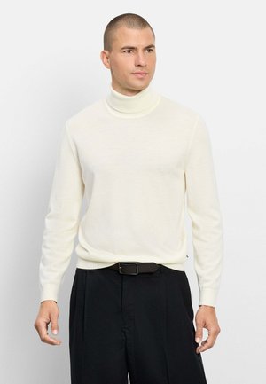 Mann, der einen weißen Rollkragenpullover und schwarze Hose trägt, steht vor einem schlichten weißen Hintergrund und schaut zur Seite.