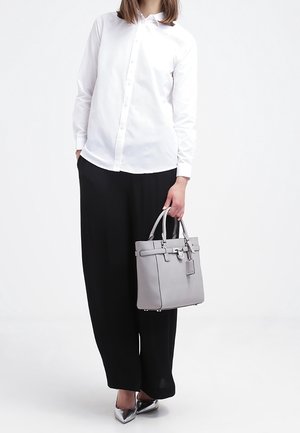 Grå læderhåndtaske med et struktureret design, der har en aftagelig mærkat og sølvfarvet hardware, båret af en person i hvid skjorte og sorte bukser.