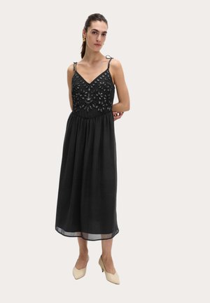 Donna che indossa un abito midi nero senza maniche con ricami floreali di perline sul corpetto e décolleté beige con punta appuntita.