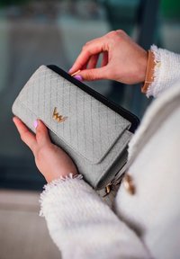 Gray wallet with a diamond quilted pattern, black zipper, and gold logo on the front. The wallet is held by a hand with lavender nails.