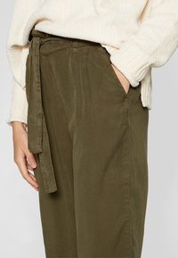 Olijfgroene broek van zacht materiaal, met een hoge taille, strikceintuur en zijzakken. Het materiaal heeft een licht gestructureerde afwerking.