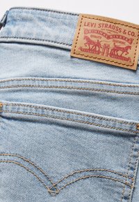 Heledu sinine denim lõuend, millel on kontrastsed kollased õmblused, ja mis sisaldab nahklabelit vöökohtadel, millele on pressitud tekst ja ikoonid.